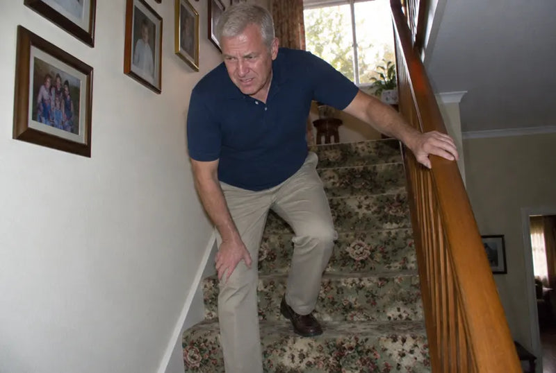 A man in his late fifties stopped partway down a staircase, one hand on the rail, the other pressed below his kneecap